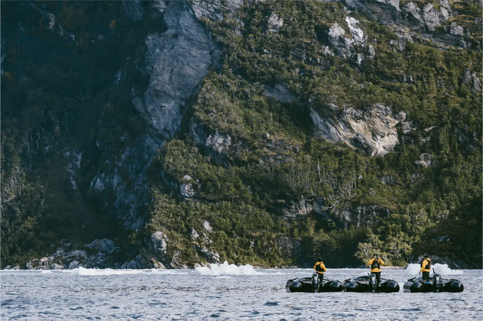 Three people in yellow jackets standing on small black motorized boats on water with a rocky, tree-covered cliff in the background.