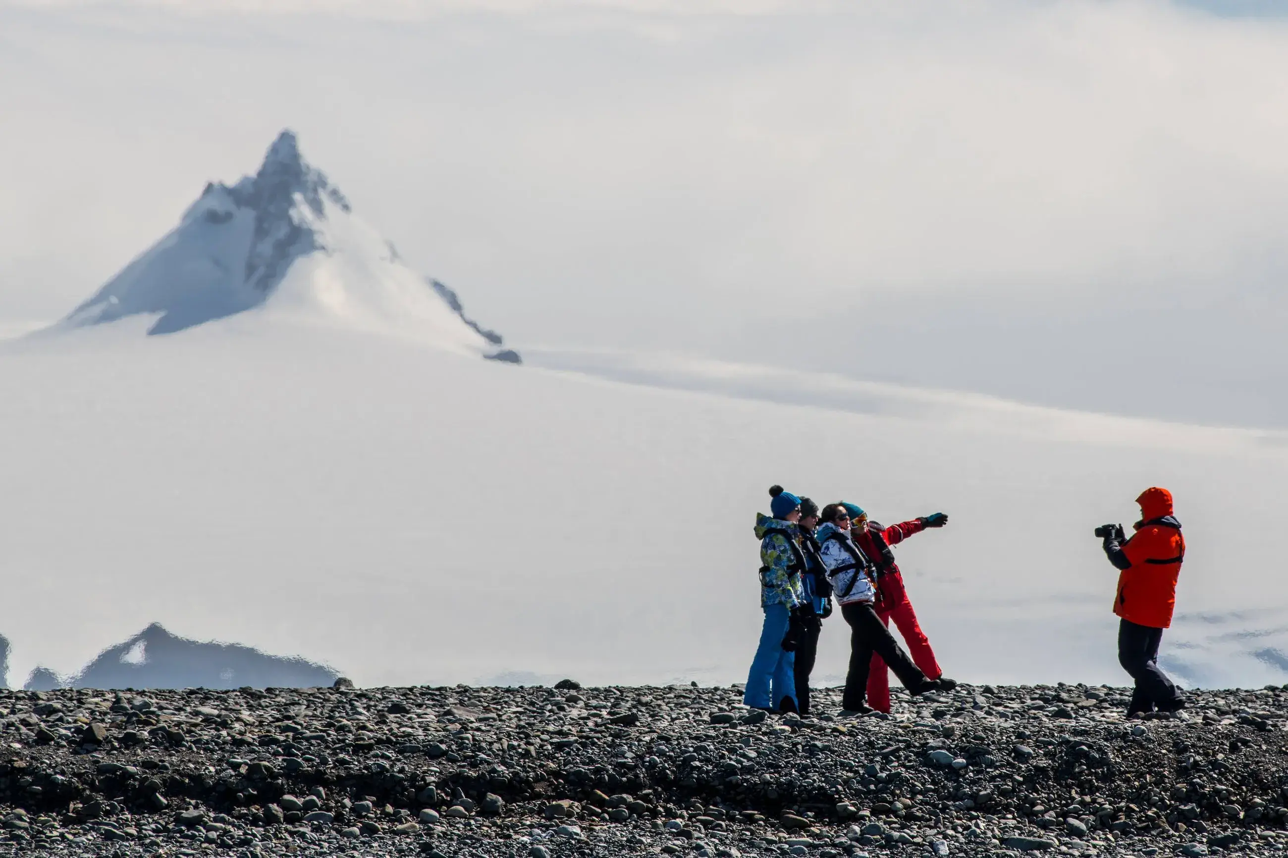 Four people in winter clothing pose for a photo on rocky terrain with a snow-covered mountain peak in the background.
