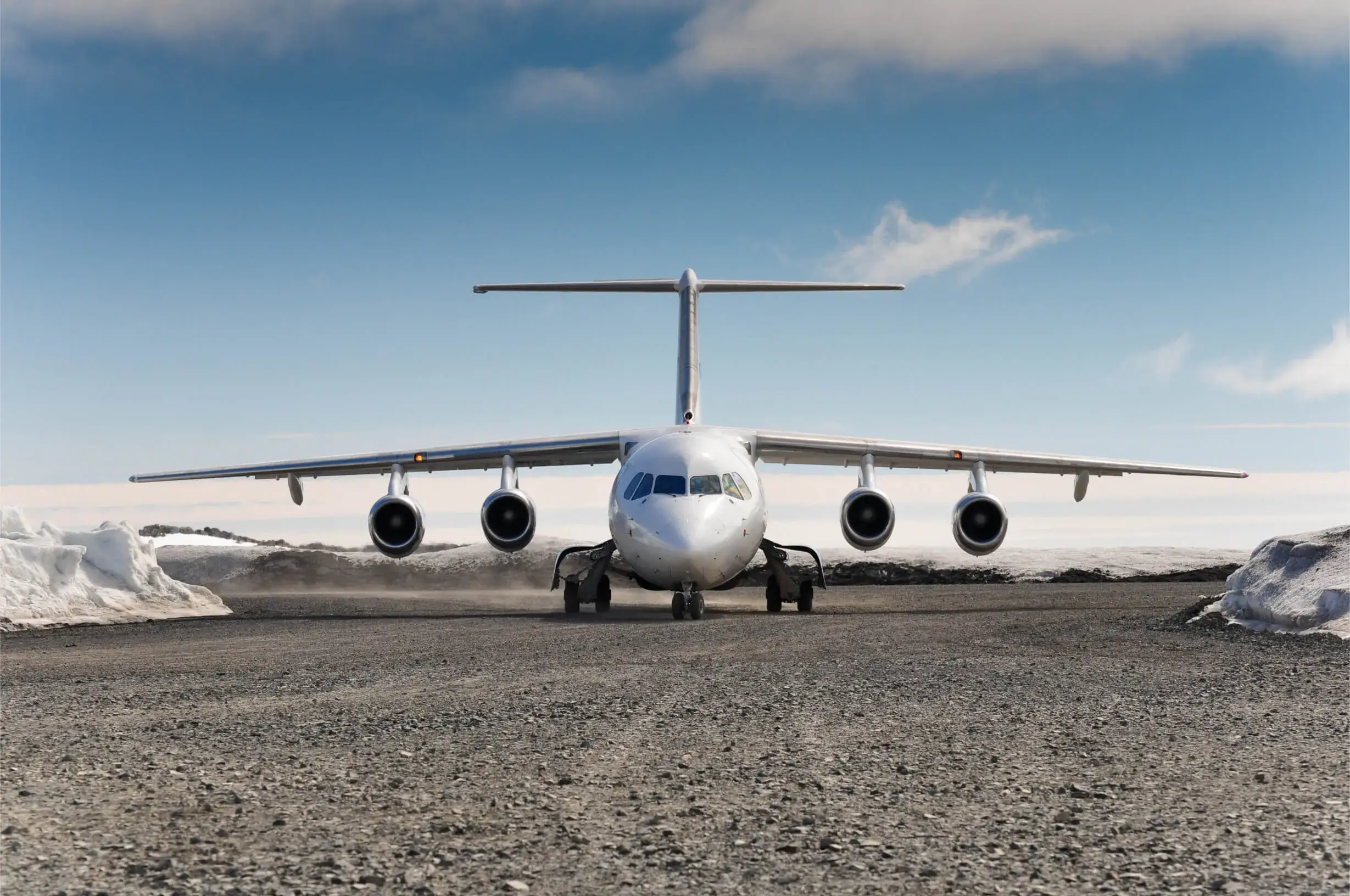 Front view of a white four-engine airplane on a gravel runway with snow on the sides and a partly cloudy sky.