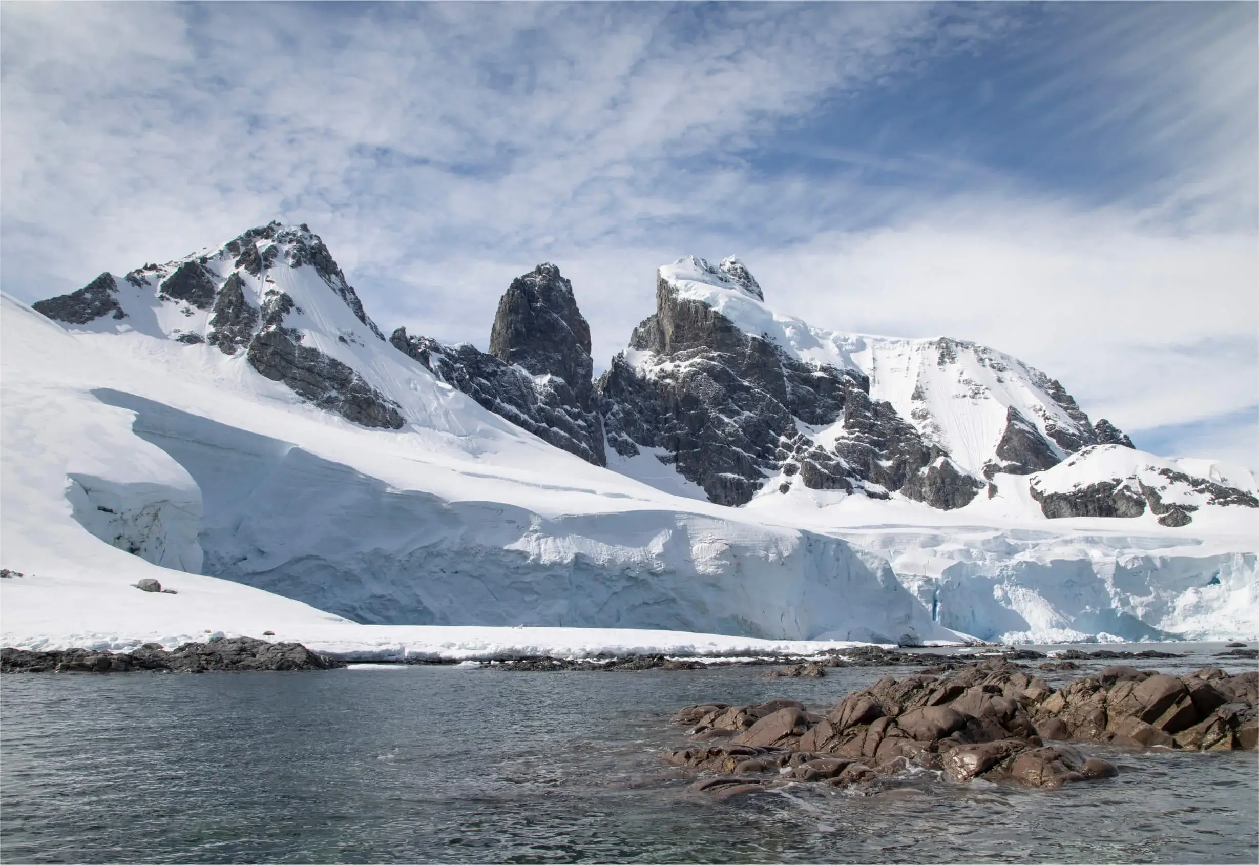 Snow-covered rugged mountain peaks behind a rocky shoreline and calm water under a partly cloudy sky.