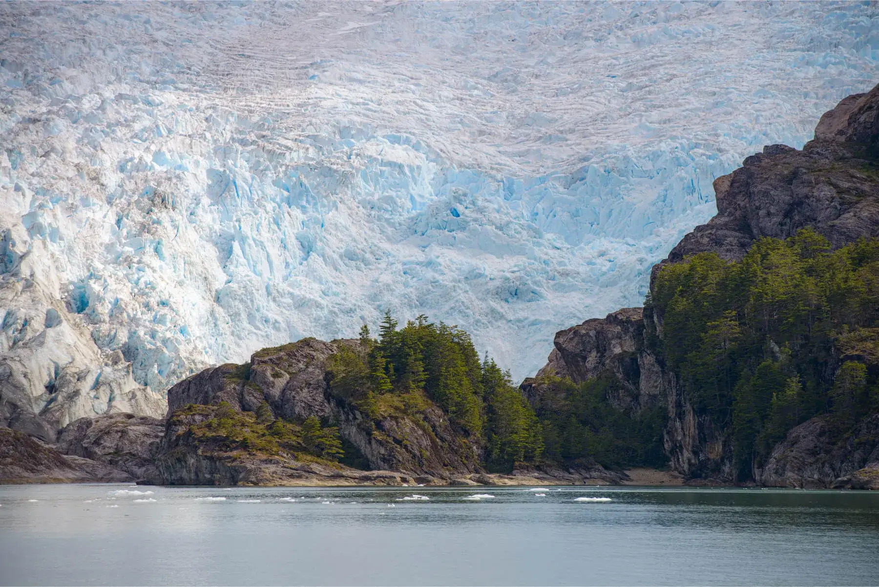 Calm water in front of rocky islands with green trees, backed by a large blue glacier.
