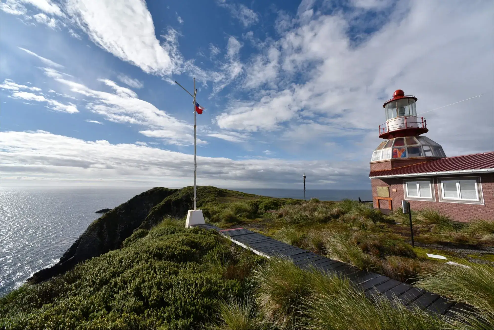 Lighthouse with red roof on grassy cliff by the ocean under a partly cloudy sky.