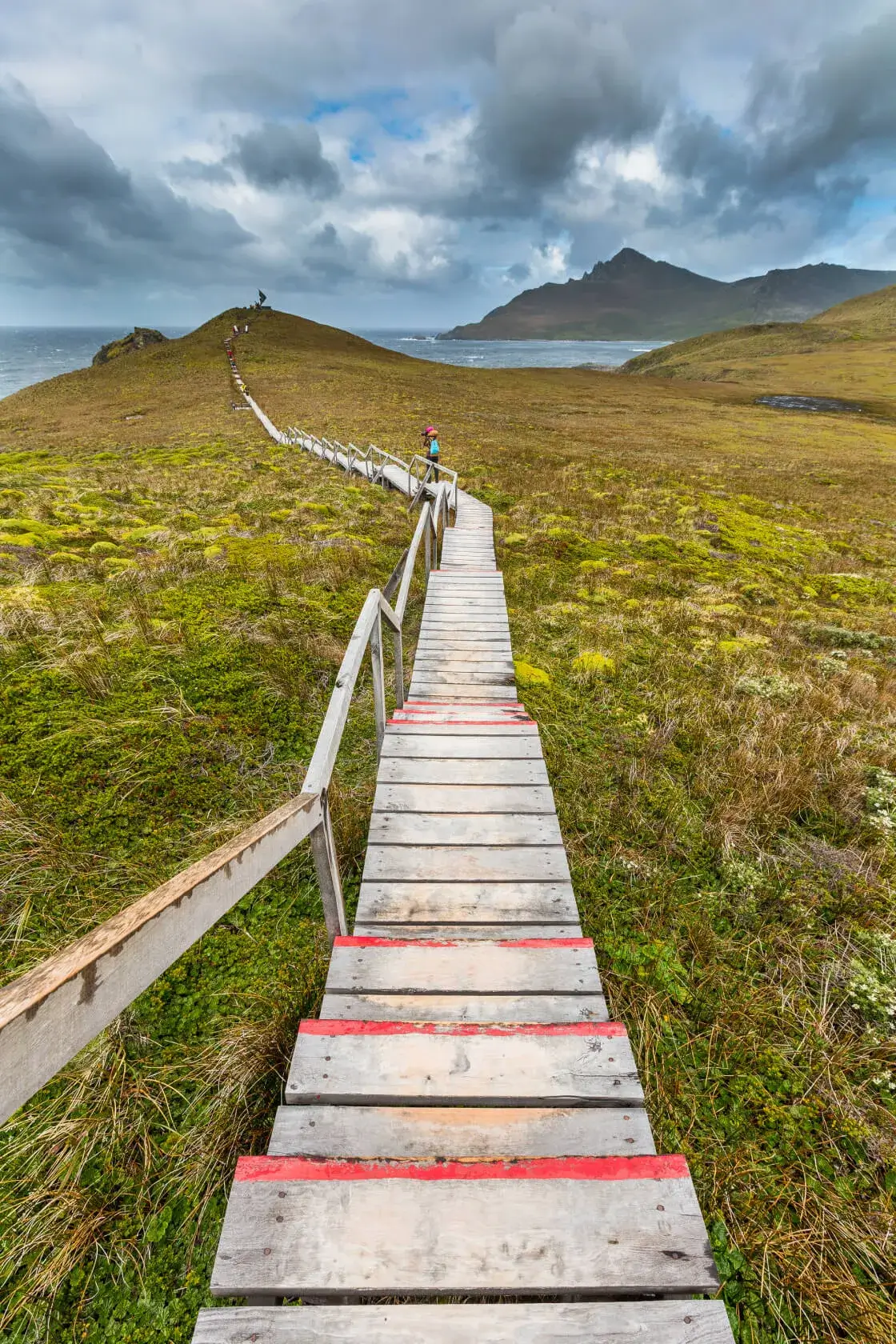 Wooden boardwalk with red markings winding through a grassy landscape toward a hill, with a person in a pink hat standing midway and cloudy sky overhead.