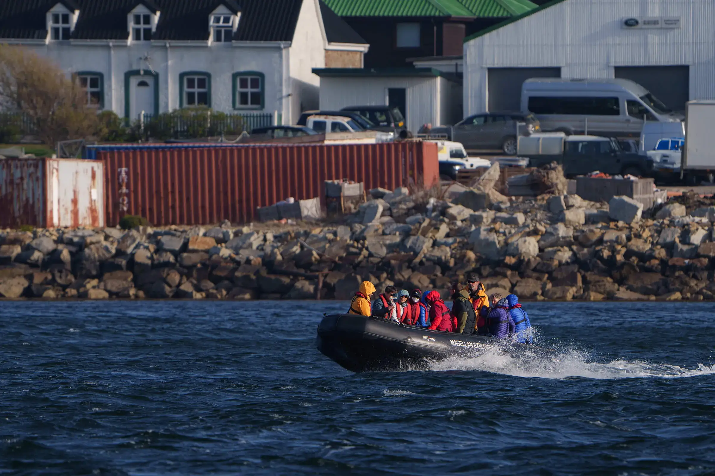 Group of people wearing colorful jackets riding a black inflatable motorboat near a rocky shoreline with buildings and parked vehicles in the background.