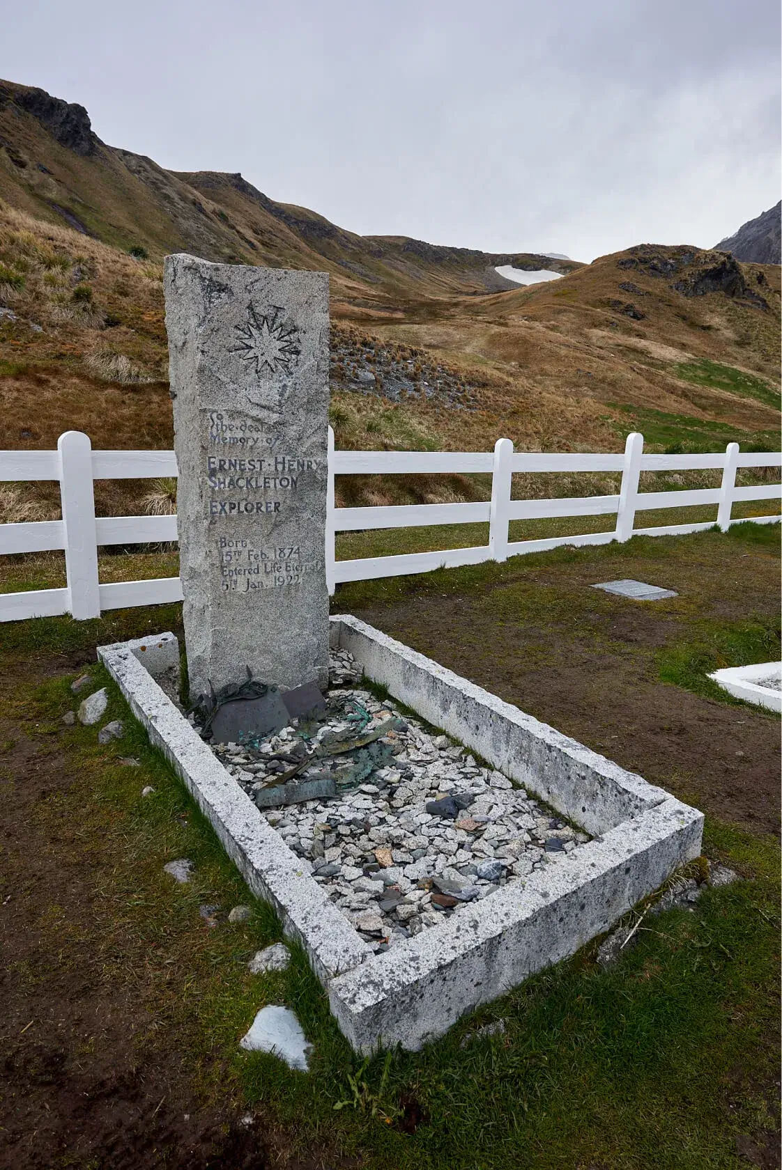 Gravestone of explorer Ernest Henry Shackleton surrounded by white fence with grassy hills in the background.