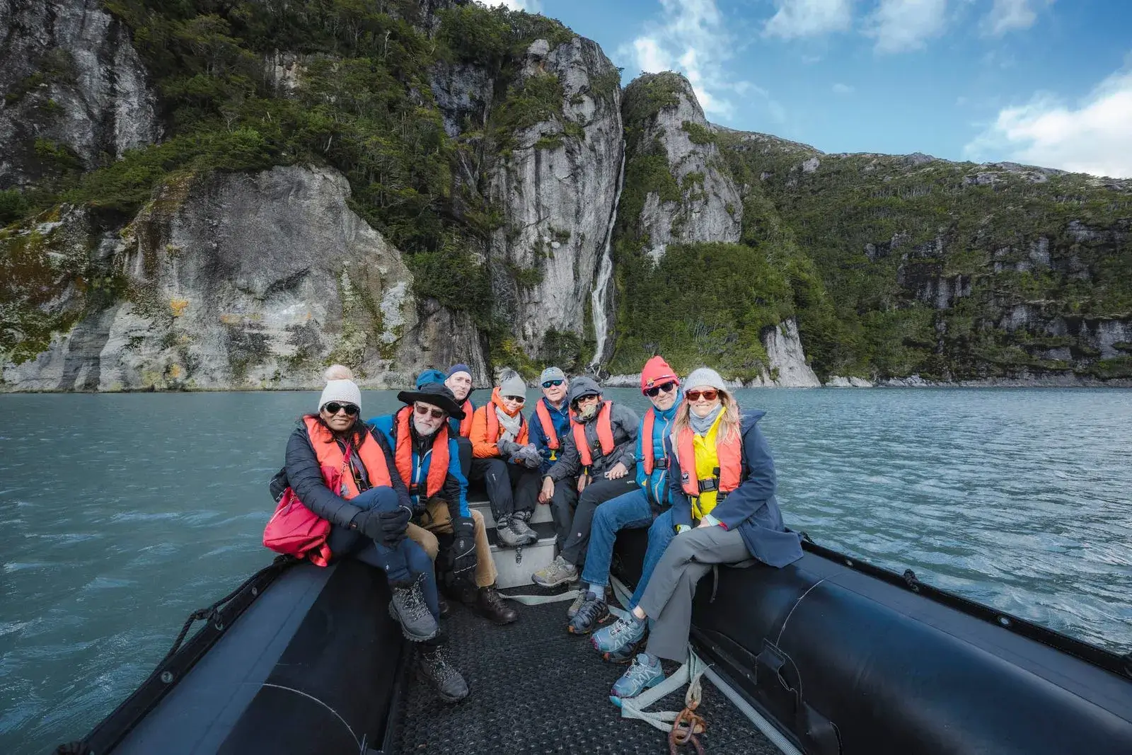 Group of eight people wearing life jackets and winter clothes sitting in a black inflatable boat on blue water with rocky cliffs and a waterfall in the background.