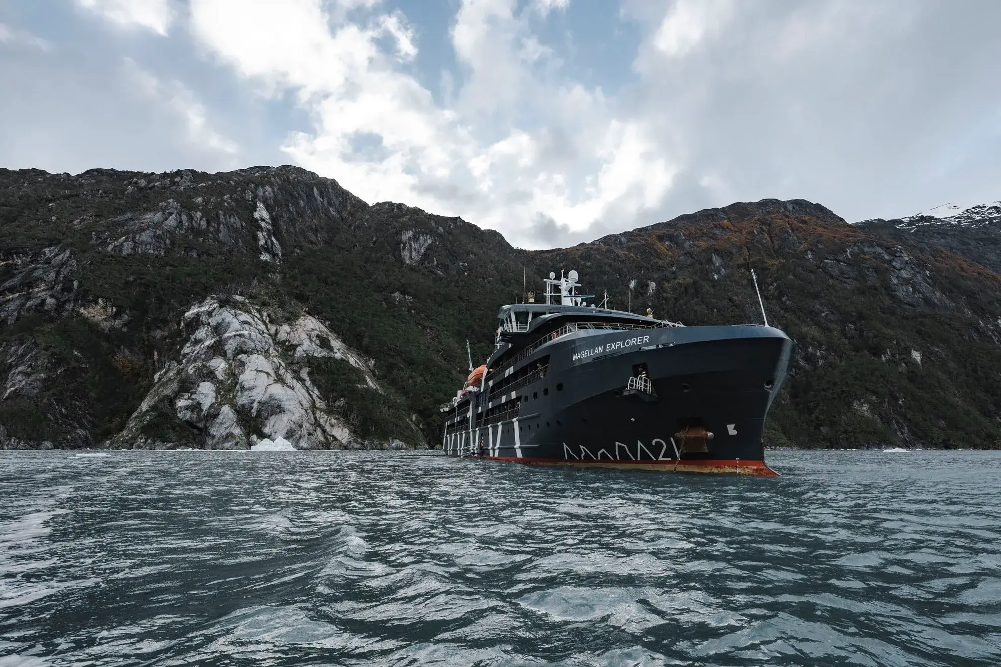 Large black ship named Magellan Explorer sailing on water with rocky, forested mountains in the background under a cloudy sky.