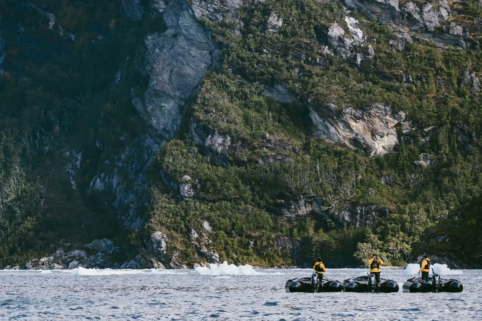 Three people in yellow jackets standing on small black inflatable boats in a body of water with a large rocky, forested cliff in the background.