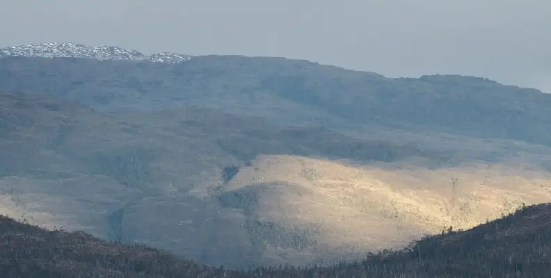 Mountain landscape with a snow-capped peak and sunlight illuminating a valley area under a cloudy sky.