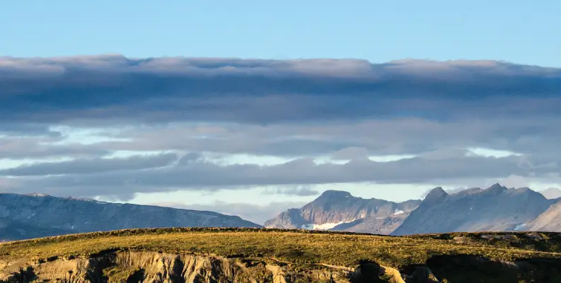 Sunlit rocky hillside in foreground with a mountain range and partly cloudy blue sky in the background.