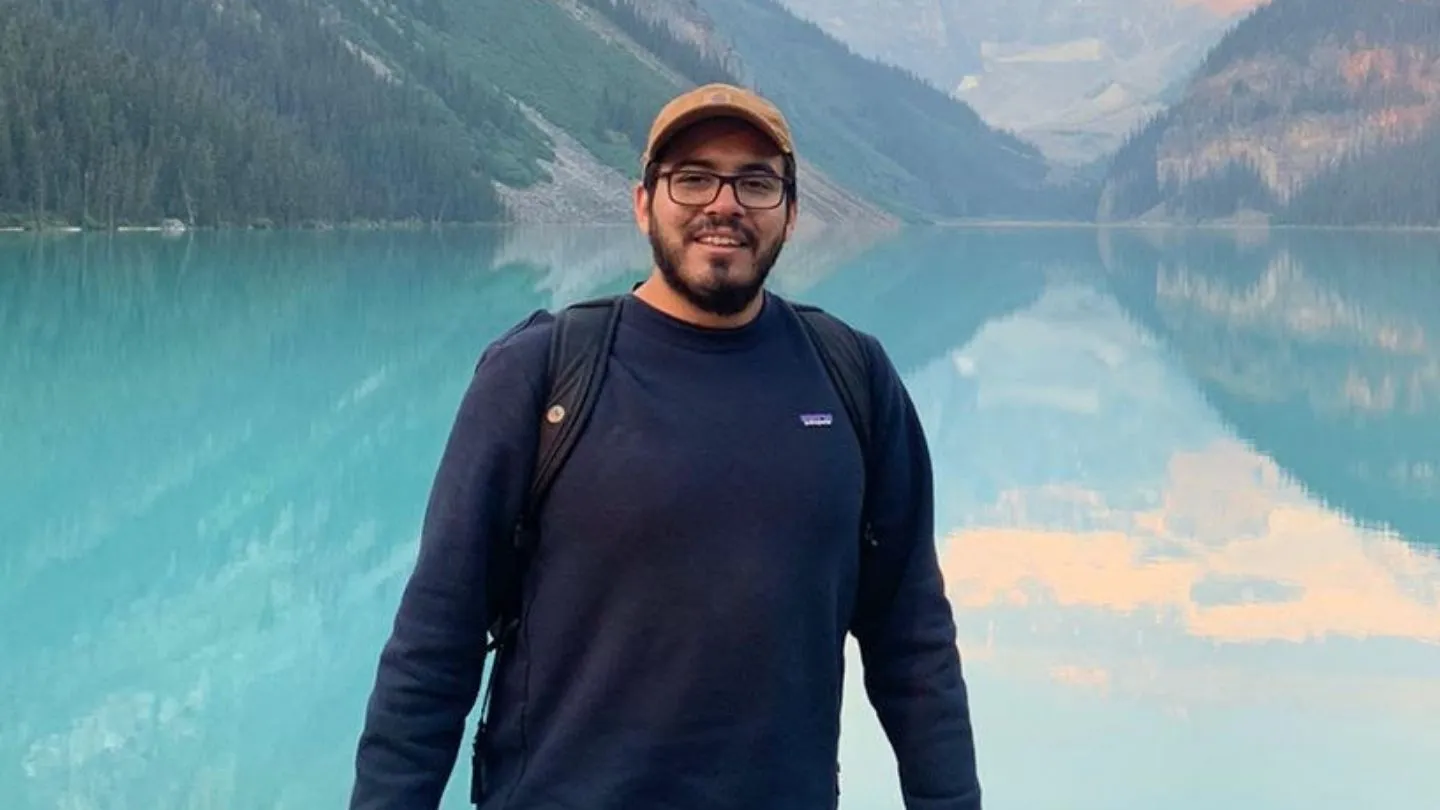 Man with glasses, beard, and brown cap smiling in front of a turquoise lake surrounded by mountains and forest.