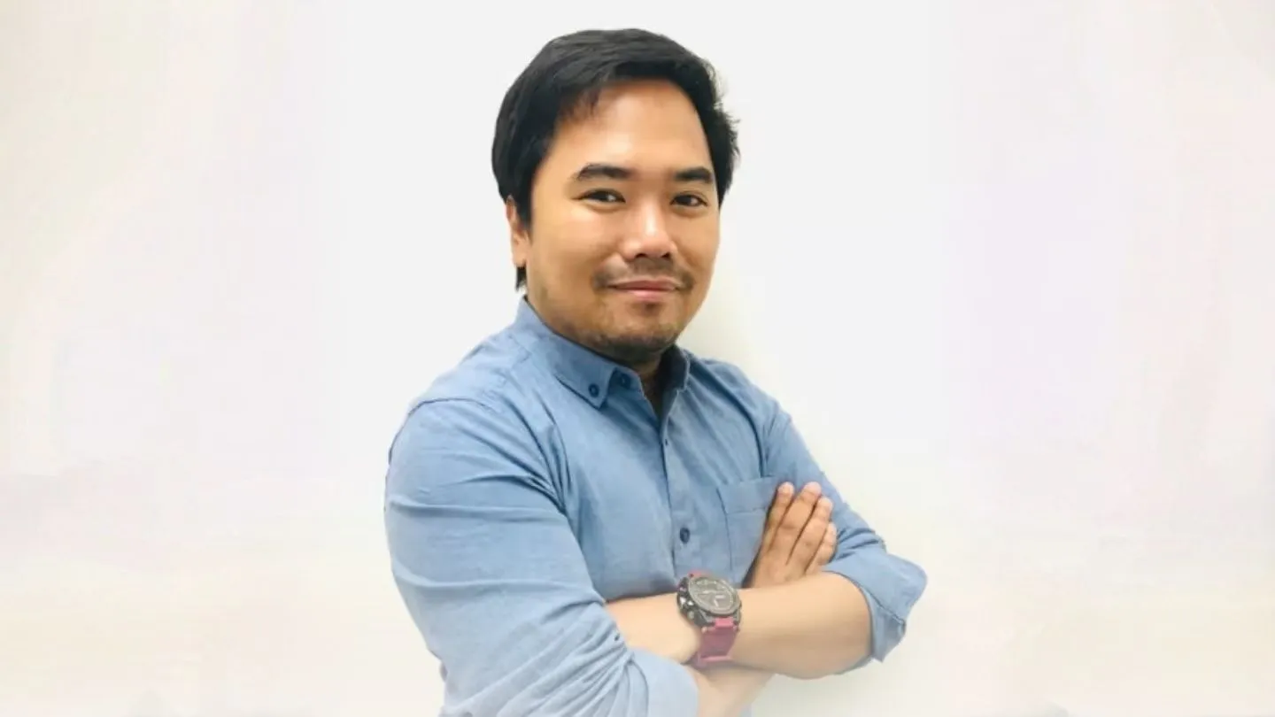 Man with folded arms wearing a blue button-up shirt and a wristwatch, standing against a plain white background.