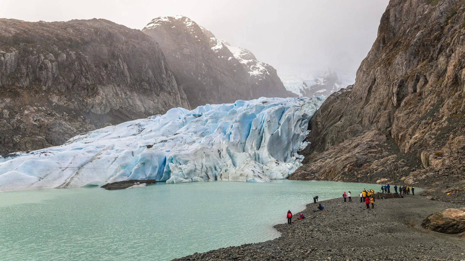 Group of people in colorful jackets standing on a rocky shore by a milky turquoise glacial lake with a large blue glacier and rugged mountains in the background.