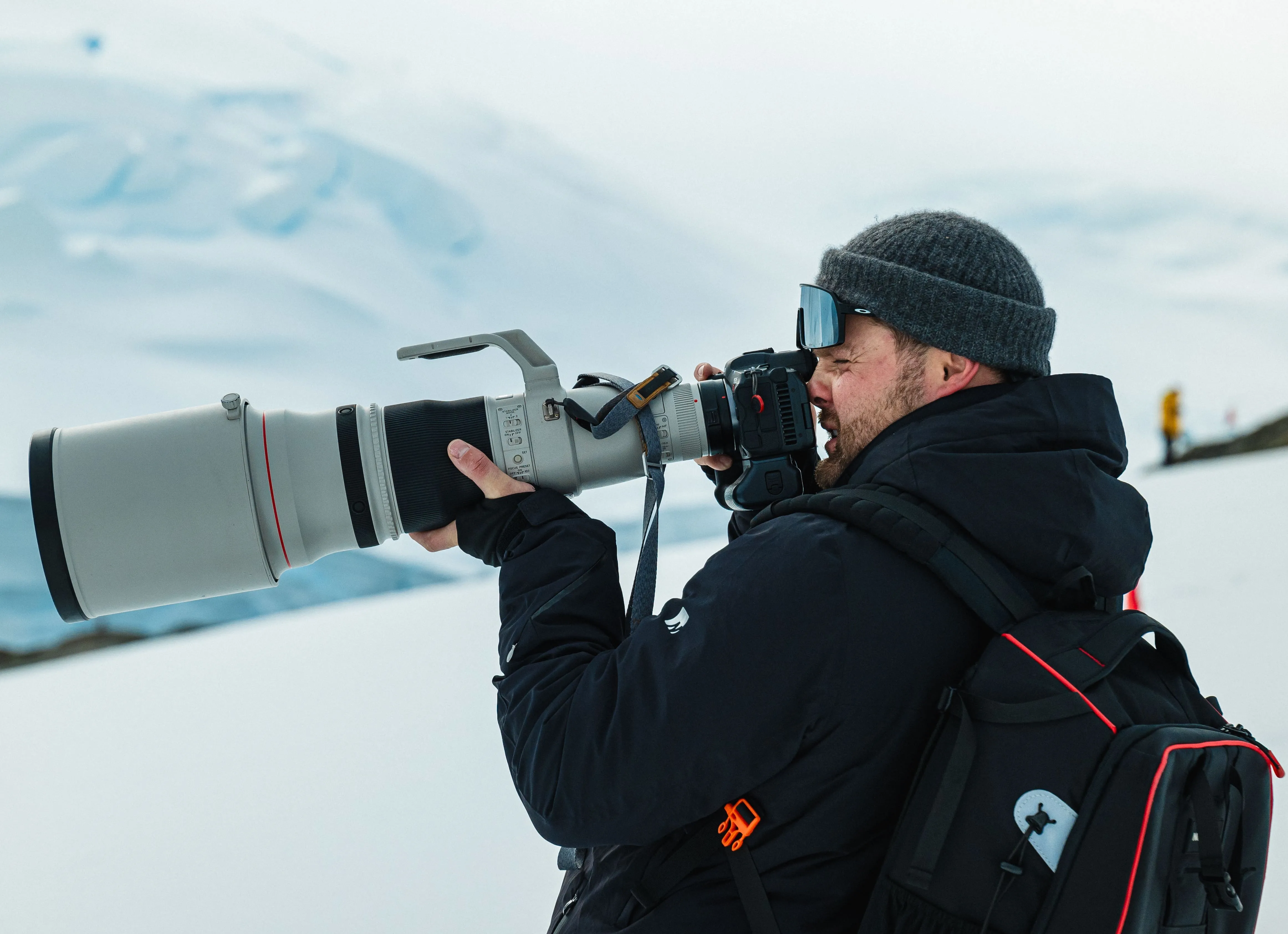 Photographer wearing winter jacket and beanie using a camera with a large telephoto lens in a snowy mountainous area.