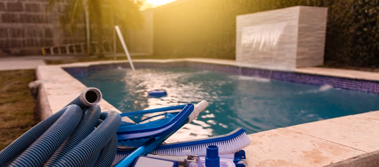 Poolreinigungsgeräte, darunter Schläuche und Bürsten, liegen am Beckenrand; im Hintergrund sind ein Pool und ein Wasserspiel bei warmem Sonnenlicht zu sehen.