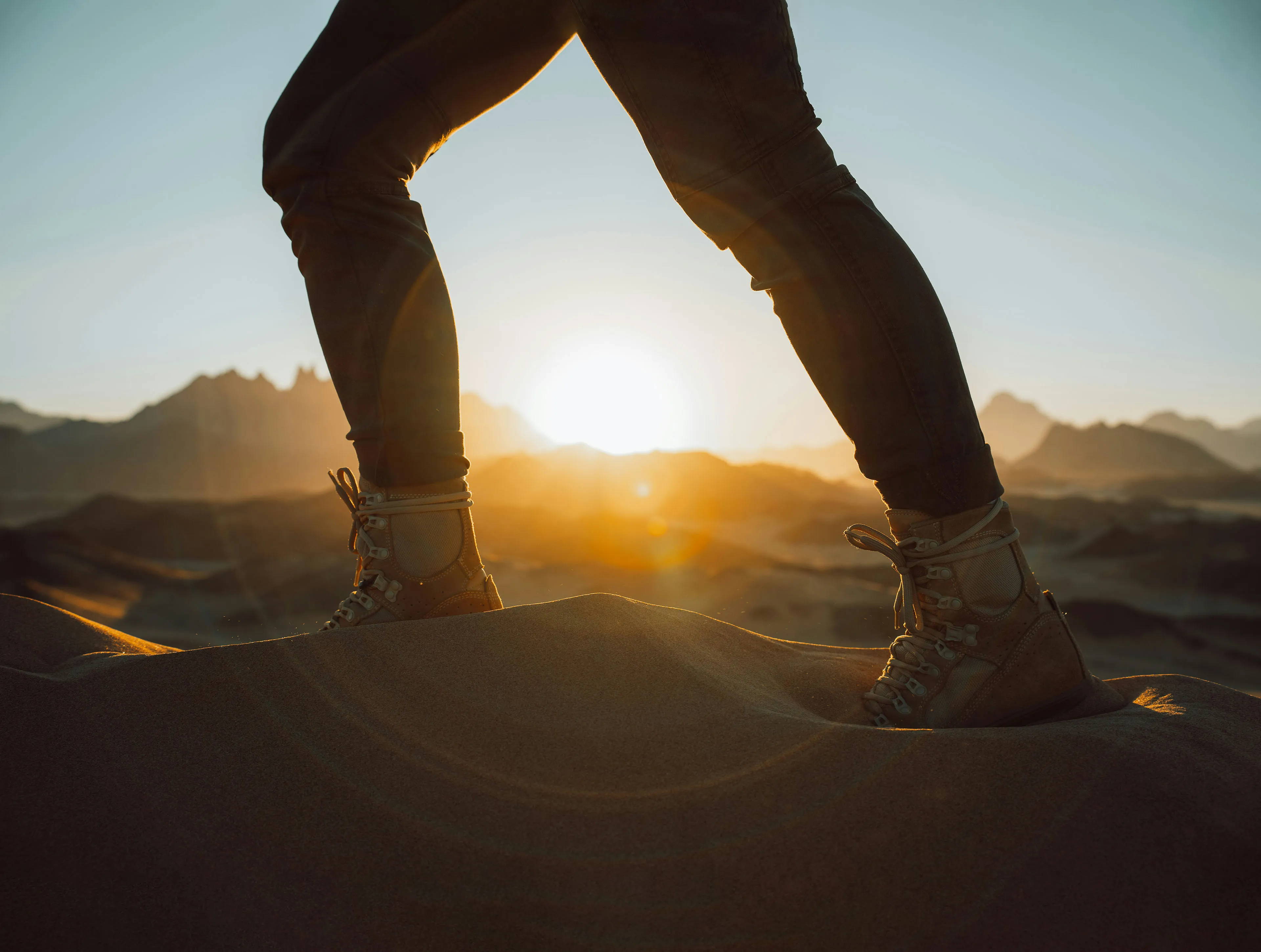 A hero section photo. The photo shows the lower legs and feet of a person wearing hiking boots and pants, standing on a sandy dune. The sun is low on the horizon, casting a warm, golden light and creating long shadows. In the background, blurry mountain silhouettes are visible against the bright sky. The focus is on the person's feet planted firmly on the textured sand.