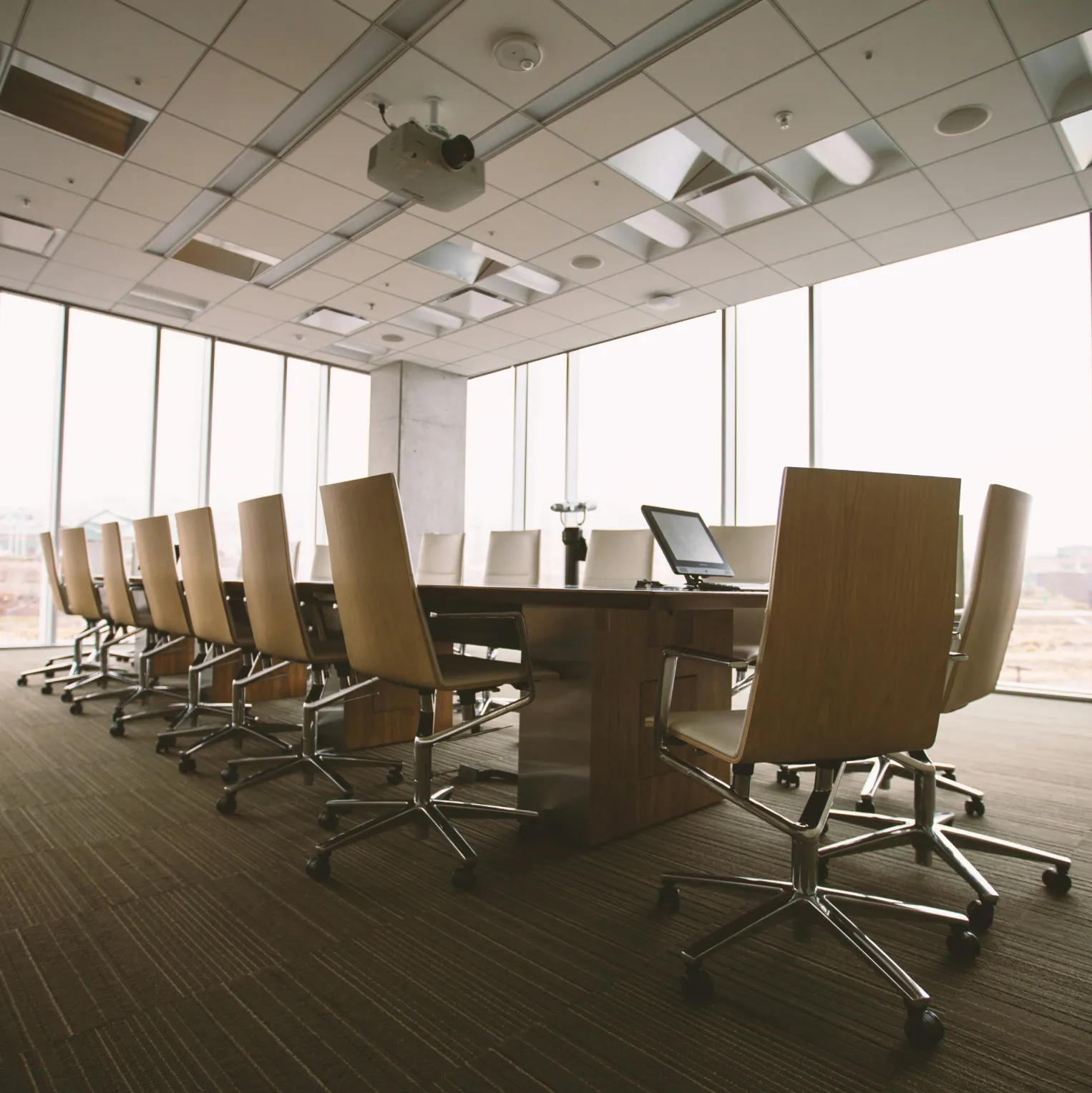 
A modern conference room is shown from a low angle, with a long, polished wooden table dominating the center of the frame. A line of sleek, light-wood chairs with chrome bases and wheels are neatly arranged on either side of the table. A laptop sits open on one end of the table, facing the chairs. The room is brightly lit, with large floor-to-ceiling windows running the length of the wall on the right, offering a slightly hazy, wide view of a distant cityscape and a light-colored sky. The floor is covered in a plush, patterned brown carpet, and a projector hangs from the ceiling, which is a grid of acoustic tiles. The overall atmosphere is one of professional, quiet readiness.