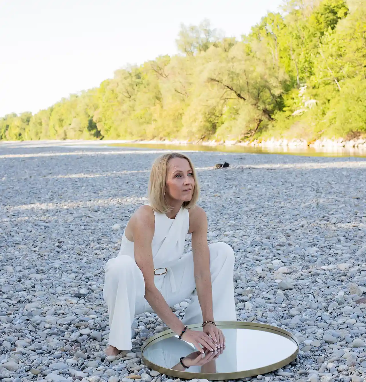 Heidi Schliebe in weißer Kleidung hockt auf einem steinigen Flussbett und berührt einen runden Spiegel. Die Szene wirkt ruhig und naturverbunden – passender Kontext zu den Raum-Harmonie-Themen.