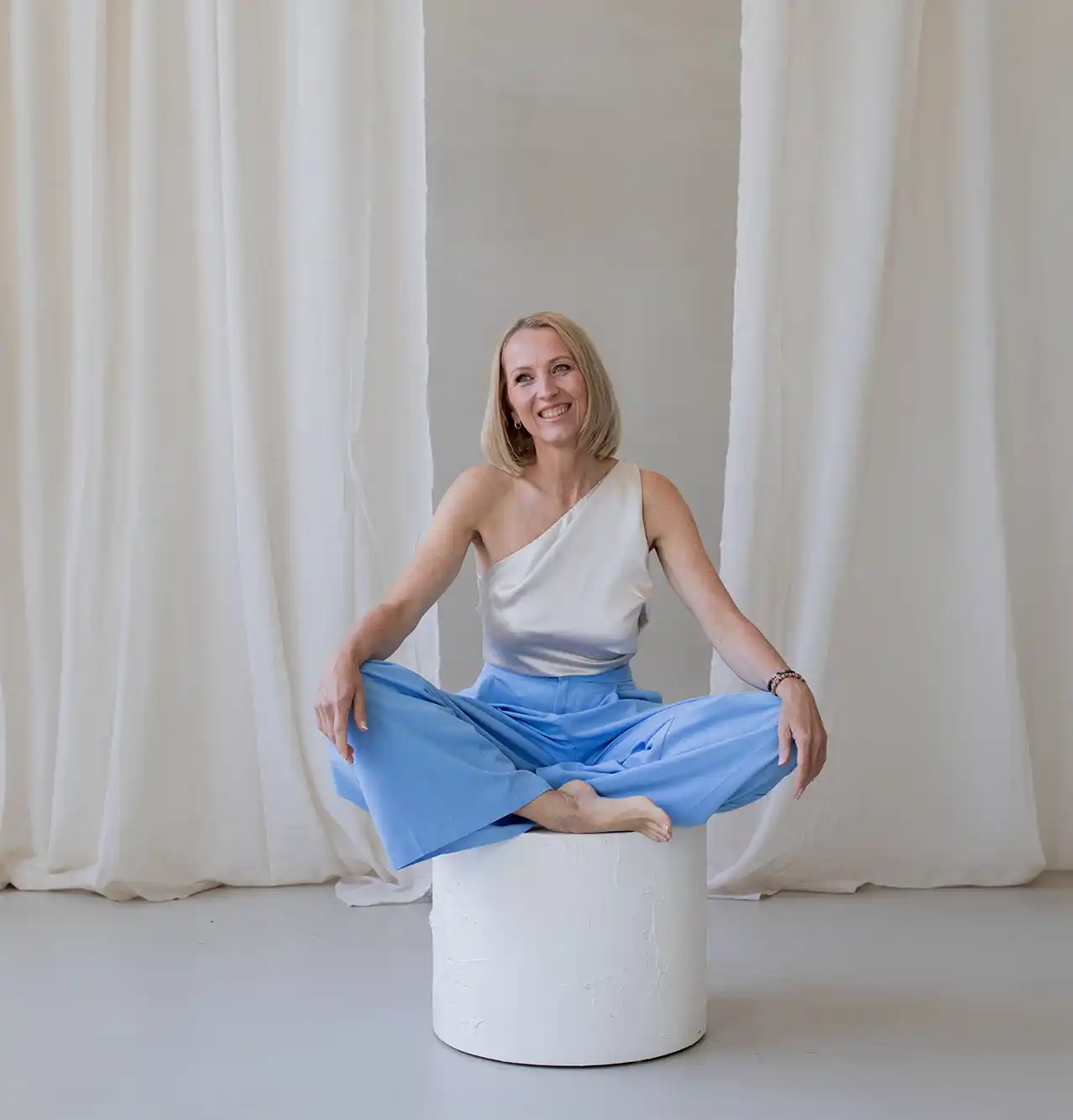 Heidi Schliebe sitzt barfuß im Schneidersitz auf einem weißen Zylinderhocker, trägt ein helles Oberteil und weite blaue Hosen und lächelt vor weißen Vorhängen.