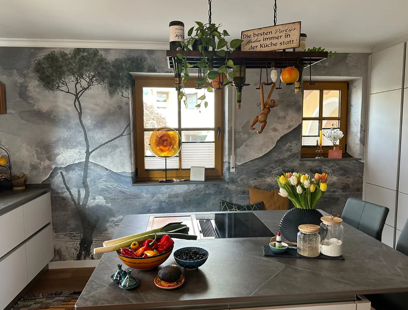 Moderne Küche mit künstlerischer Landschafts-Tapete, Kochinsel, bunten Lebensmitteln, vielen Pflanzen und warmen Holzfenstern.