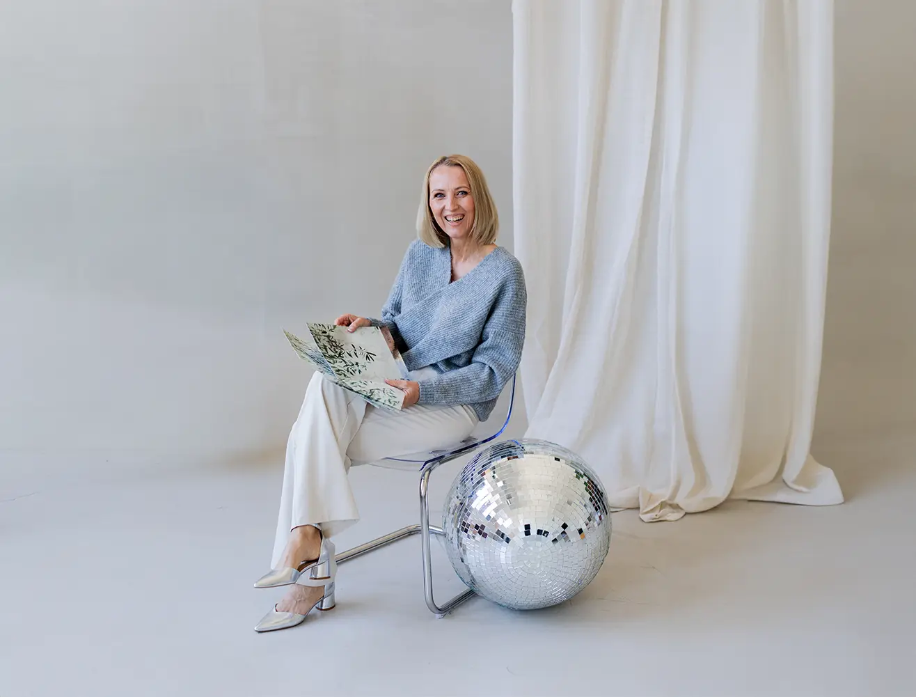 Heidi Schliebe sitzt auf einem Stuhl, hält mehrere Stoffmuster mit Pflanzenmotiven und lächelt. Neben ihr steht eine große silberne Diskokugel, im Hintergrund helle Vorhänge.