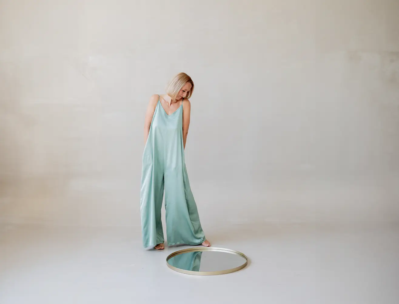Heidi Schliebe steht barfuß in einem mintgrünen, fließenden Overall und blickt auf einen runden Spiegel, der auf dem Boden vor ihr liegt.