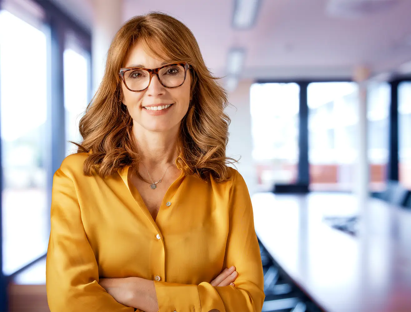 Eine Frau mit brünetten Haaren und Brille steht lächelnd in einem modernen, hellen Büro und hat die Arme verschränkt.