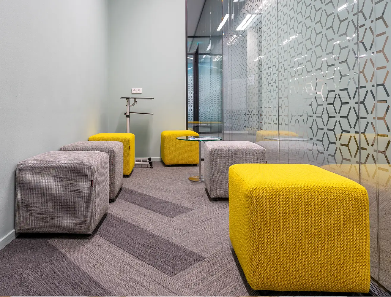 Moderner Bürobereich mit grauen und gelben Polsterwürfeln, Glaswänden mit geometrischem Muster und hellem Teppichboden.