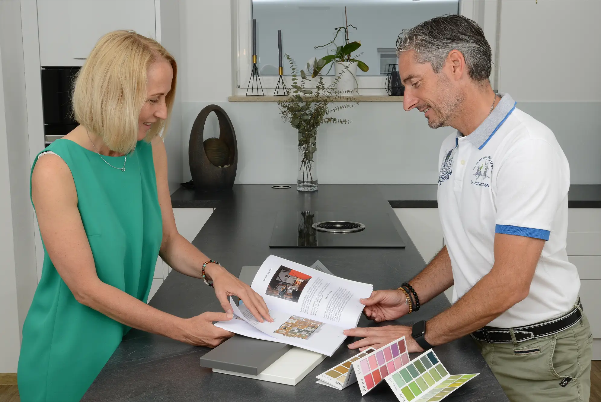 Zwei Personen stehen an einer Kücheninsel und betrachten gemeinsam ein Farb- und Materialkonzept mit Musterkarten und Unterlagen.