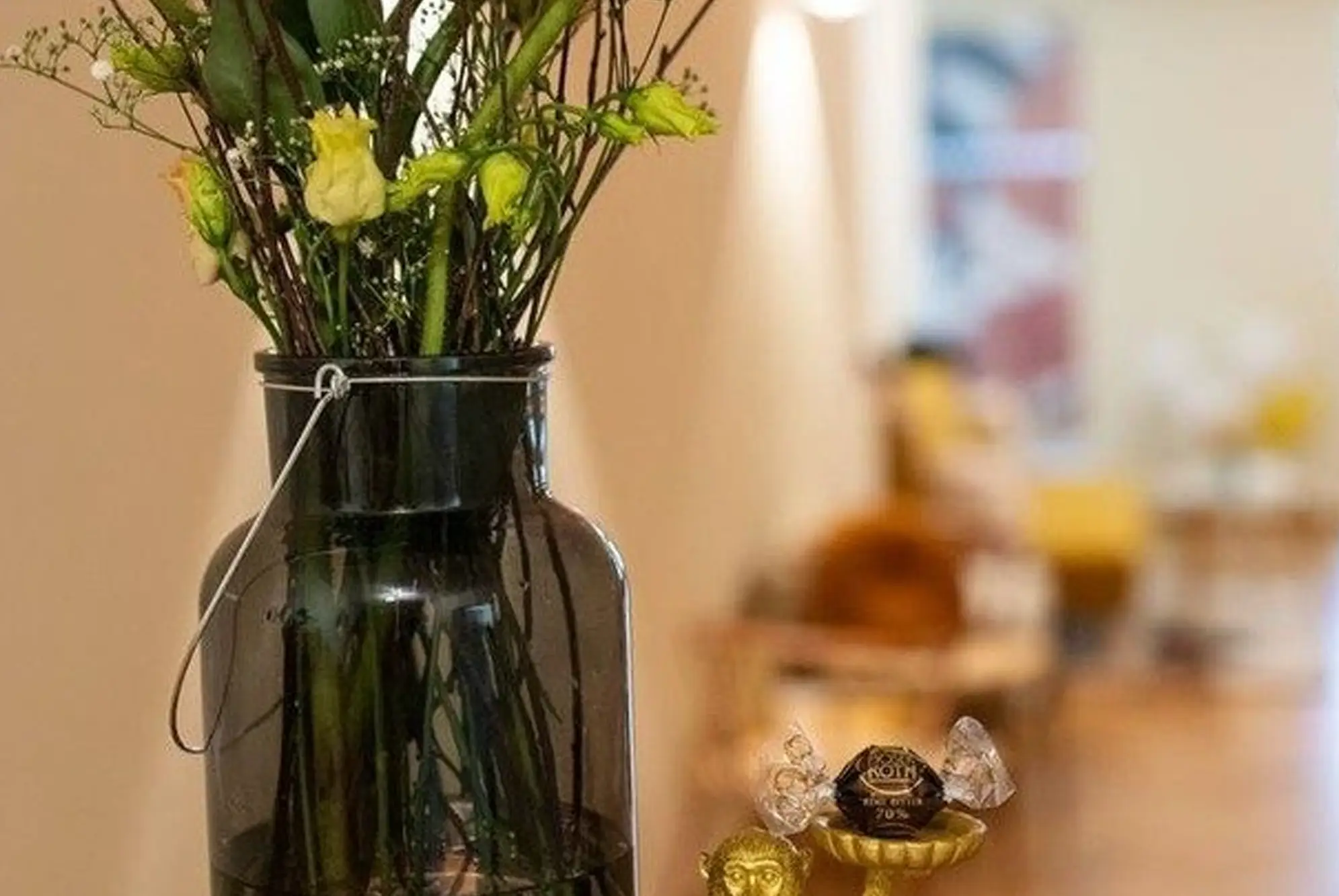 Große Glasvase mit frischen Blumen auf einer kleinen Ablage, dahinter ein warmer, einladender Flur mit Sitzgelegenheiten.