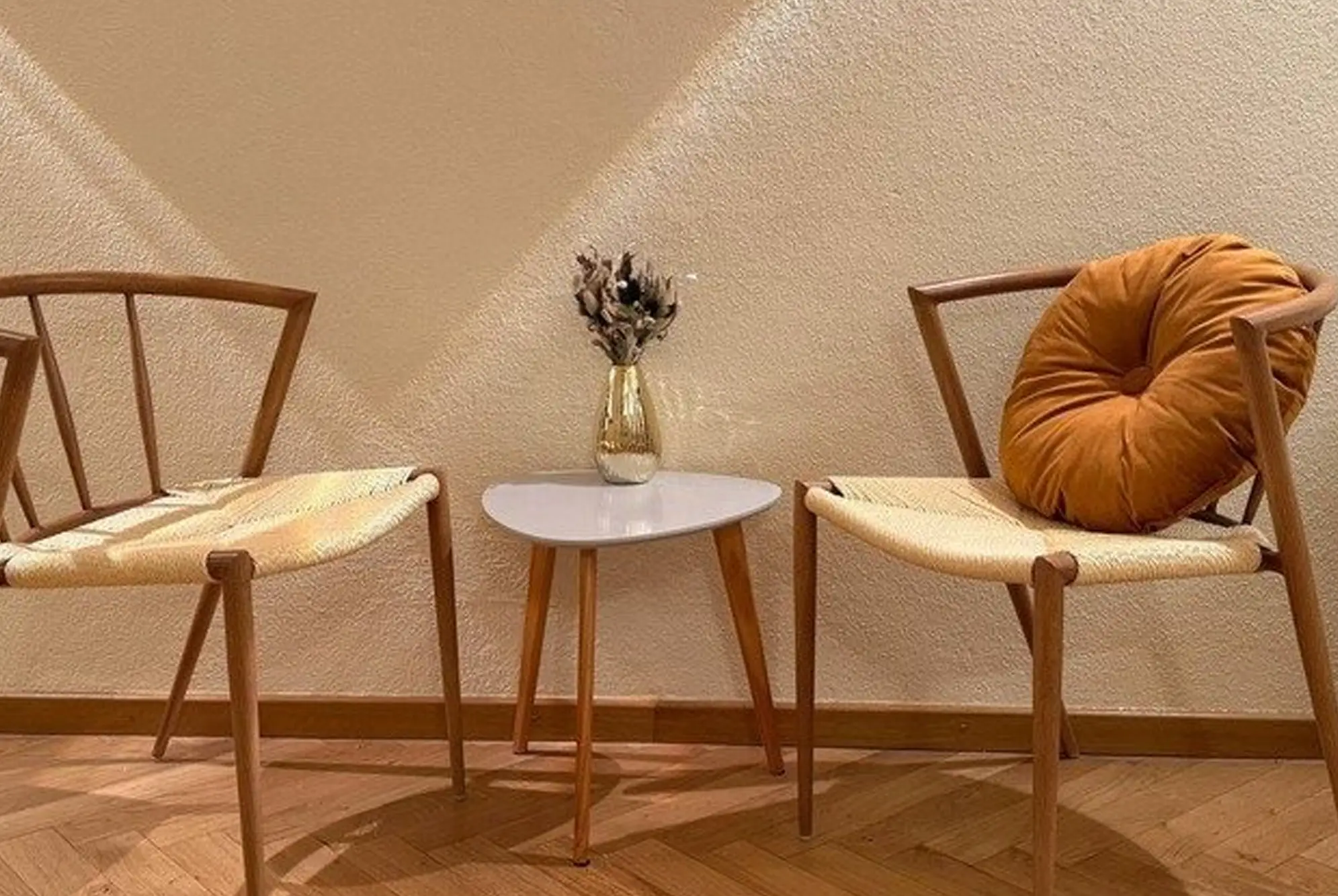Zwei Holzstühle mit geflochtenen Sitzflächen und einem kleinen weißen Tisch dazwischen, dekoriert mit einer Vase und Trockenblumen.