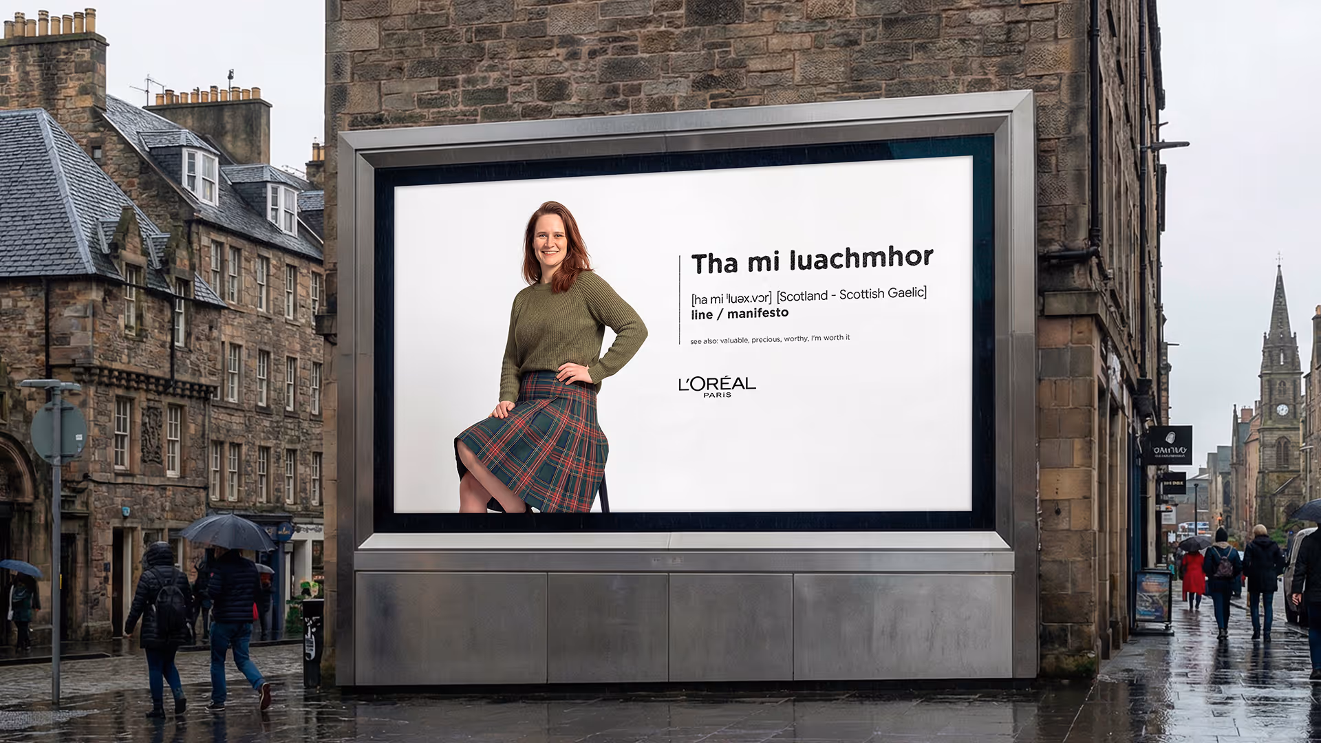 Advertising billboard on a city street showing a woman in a green sweater and plaid skirt with text in Scottish Gaelic and L'Oréal Paris logo.