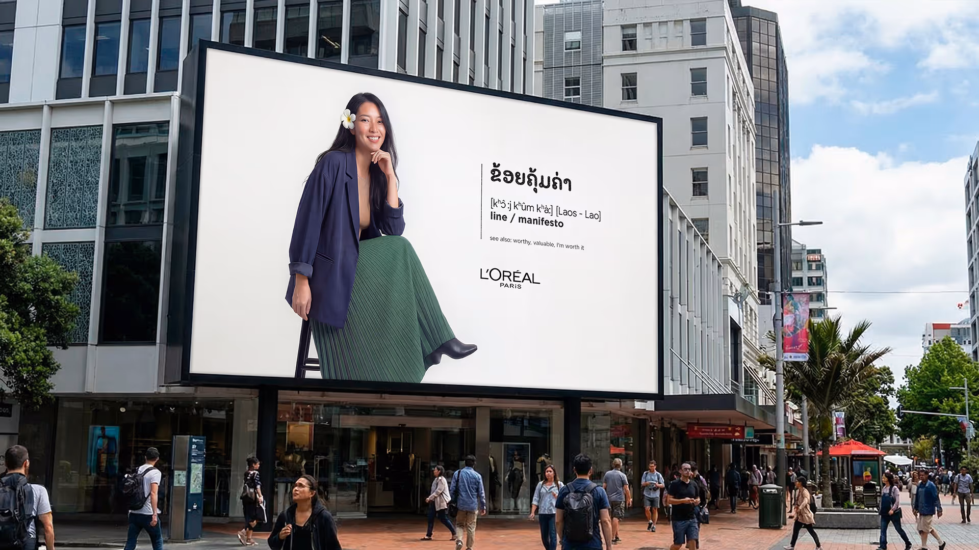 Outdoor billboard in city with a woman wearing a dark jacket and green skirt sitting on a stool next to text in Lao script and L'Oréal Paris logo.