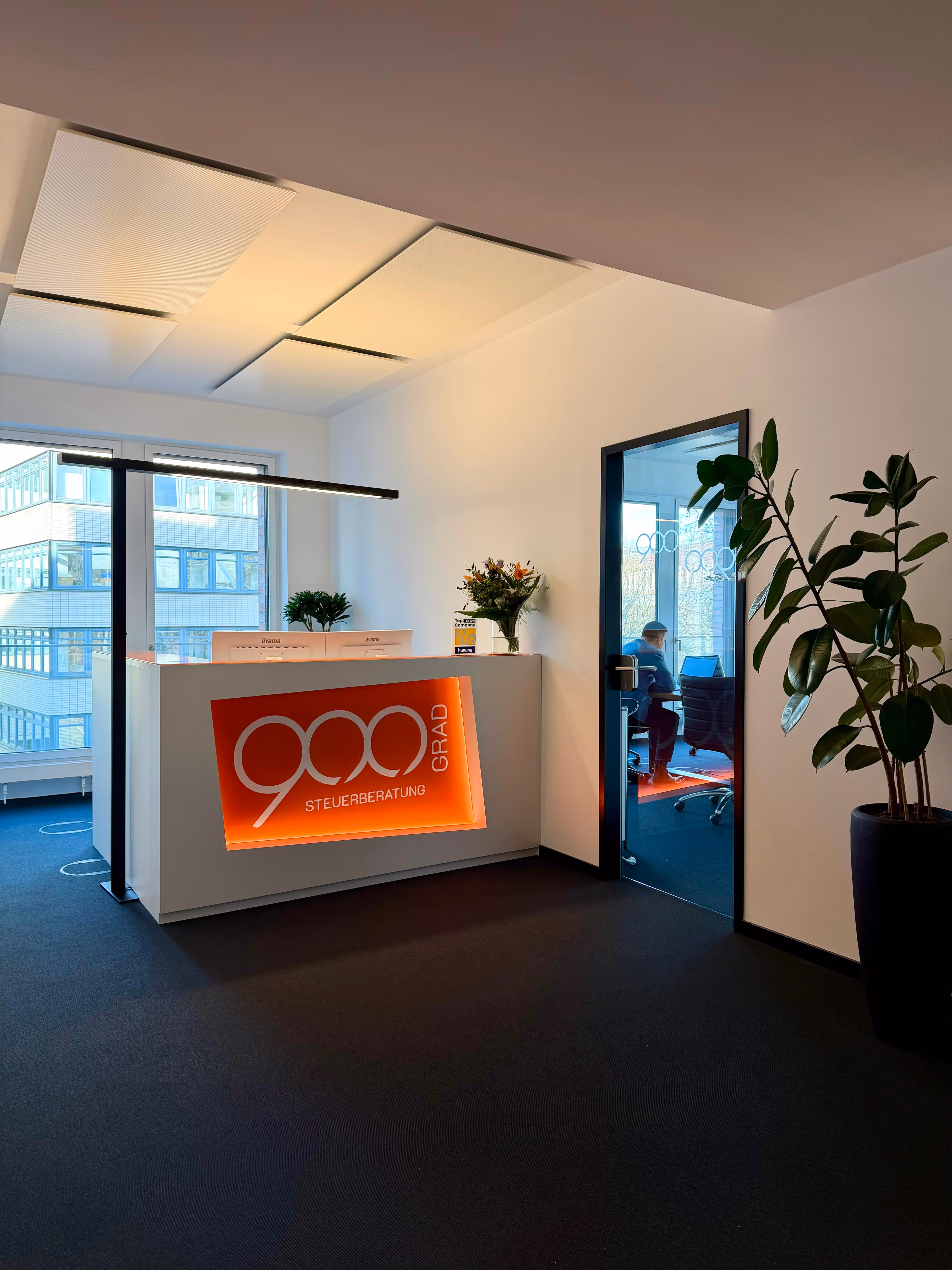 Modernes Büro mit Holzboden, schwarzen Regalen, weißen Wänden, Holzbalkendecke und einem orangefarbenen Logo an der Wand.