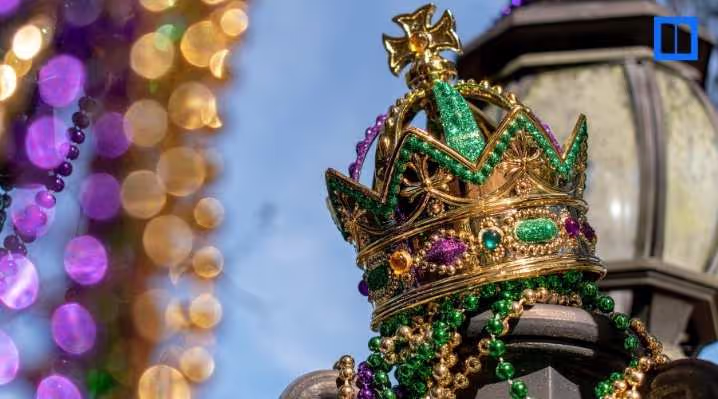 Close-up of a glittering gold, green, and purple Mardi Gras crown perched on a street lamp, adorned with colorful beads against a blurred background of festive lights.