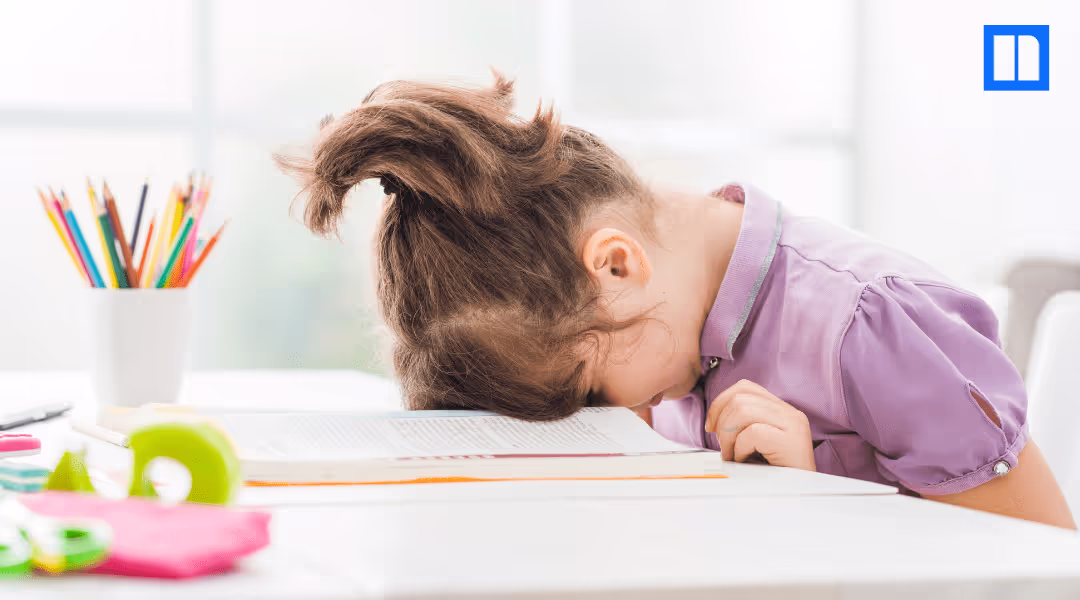 A young child with her hair in a messy ponytail is slumped over a book on a desk, appearing tired or stressed. There are school supplies like colored pencils and a pair of scissors on the desk.