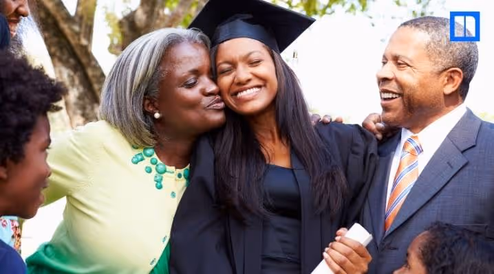 A joyful graduate in a black cap and gown being kissed on the cheek by her mother while her father and family smile during a graduation ceremony.