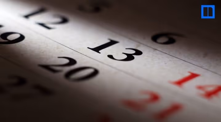A dramatic, low-angle close-up of a paper calendar with a spotlight focusing on the number 13. The surrounding dates are blurred and in shadow, and a small blue Newsela logo is visible in the top right corner.
