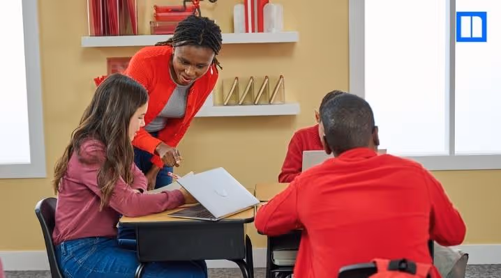 "A female teacher in a red sweater leans over to help a female student working on a laptop at a desk, with other students in the classroom. The Newsela logo is in the top right corner."