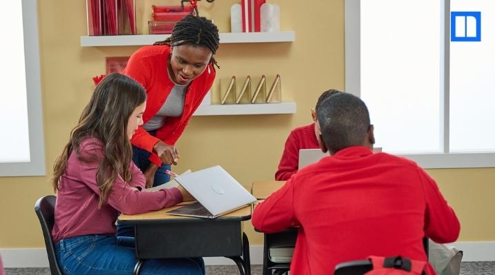 "A female teacher in a red sweater leans over to help a female student working on a laptop at a desk, with other students in the classroom. The Newsela logo is in the top right corner."