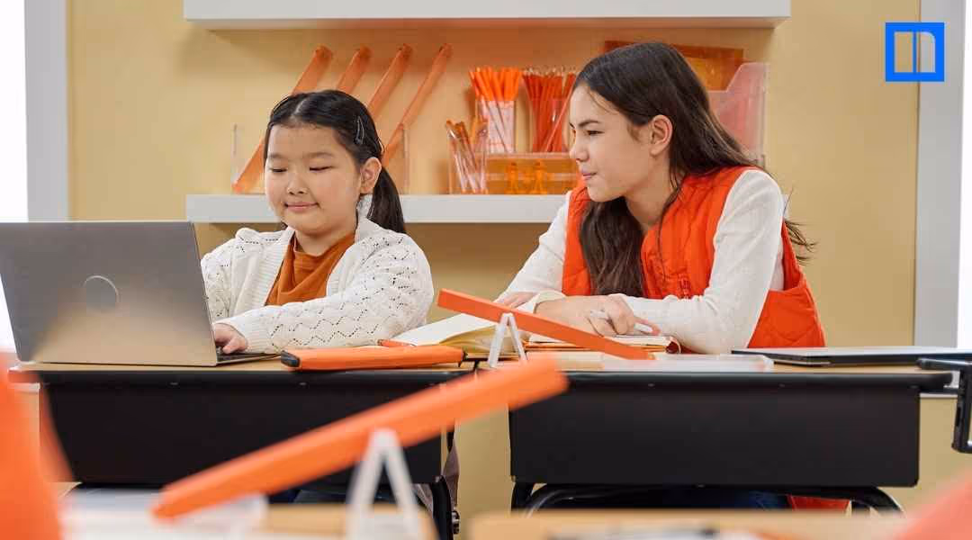 "Two female students, one using a laptop and the other observing, are seated at a desk with an orange experiment setup. The image depicts collaborative learning and a hands-on science project in a classroom setting, perfect for a blog post on the scientifi
