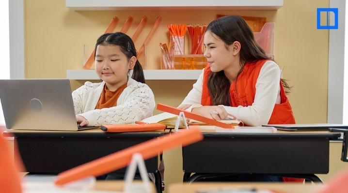 "Two female students, one using a laptop and the other observing, are seated at a desk with an orange experiment setup. The image depicts collaborative learning and a hands-on science project in a classroom setting, perfect for a blog post on the scientifi