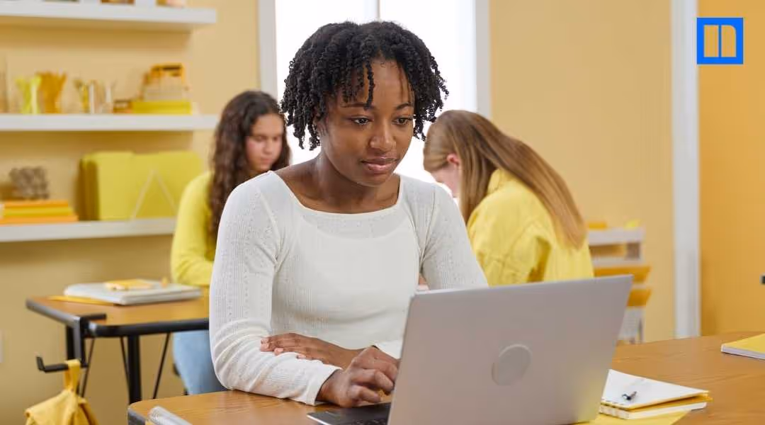 "A close-up of a focused African American female student working on a laptop at her classroom desk. Other students are blurred in the background. This image is a thumbnail for a blog post about interim assessments, representing a classroom setting where st
