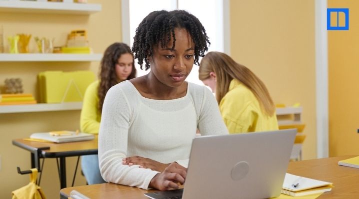 "A close-up of a focused African American female student working on a laptop at her classroom desk. Other students are blurred in the background. This image is a thumbnail for a blog post about interim assessments, representing a classroom setting where st