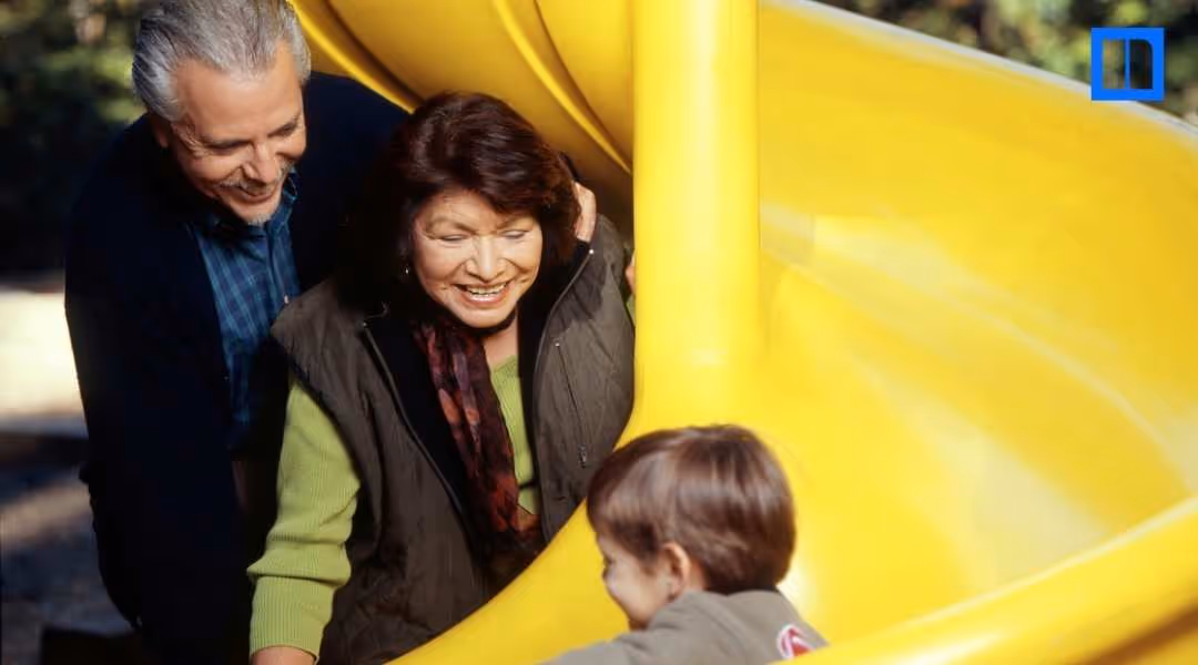 "A close-up image of a grandmother and grandfather smiling as they play with their grandchild on a bright yellow playground slide. This is a thumbnail for a blog post about Grandparents Day activities, highlighting family connections and celebratory educat