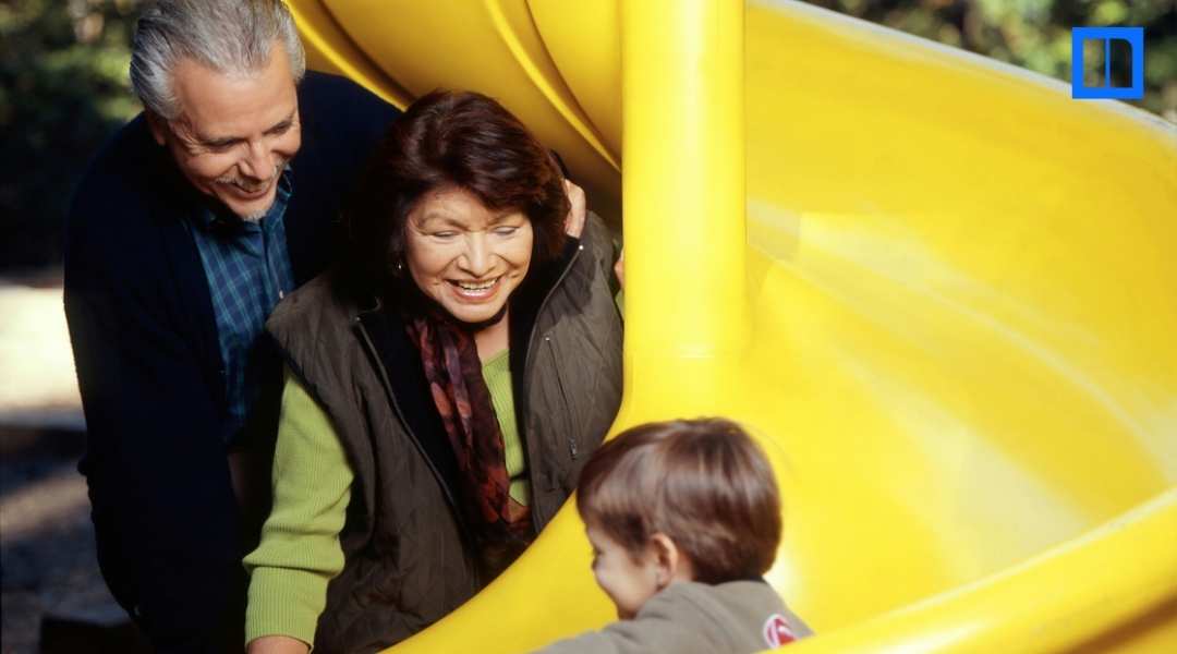 "A close-up image of a grandmother and grandfather smiling as they play with their grandchild on a bright yellow playground slide. This is a thumbnail for a blog post about Grandparents Day activities, highlighting family connections and celebratory educat