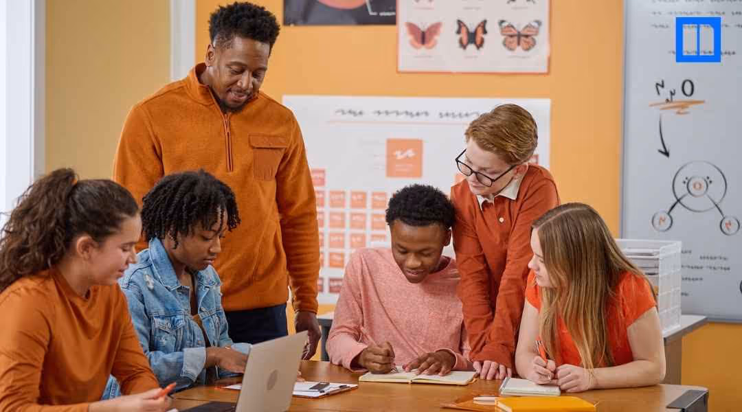 A diverse group of middle school students and their teacher collaborating on a project. The teacher is standing behind them, smiling, while the students are gathered around a table with a laptop, notebooks, and pencils, actively engaged in their work.