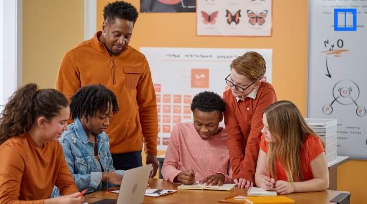 A diverse group of middle school students and their teacher collaborating on a project. The teacher is standing behind them, smiling, while the students are gathered around a table with a laptop, notebooks, and pencils, actively engaged in their work.