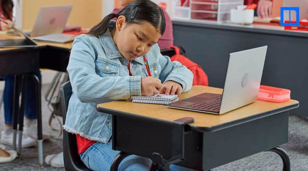 A young student with a denim jacket sitting at a desk, focused on writing in a notebook. A laptop is open on the desk next to her, and other students are visible in the background, suggesting a classroom environment.