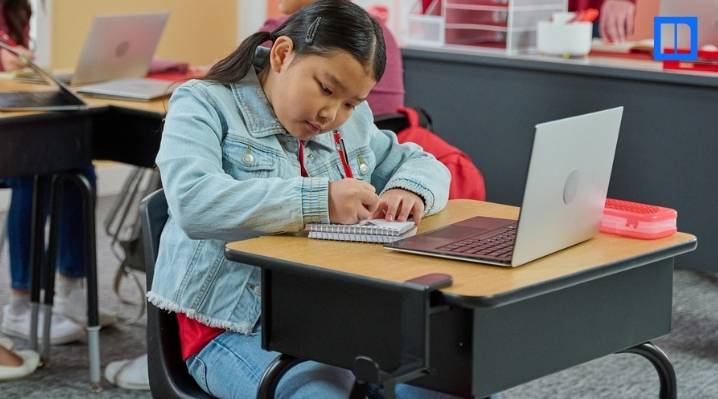 A young student with a denim jacket sitting at a desk, focused on writing in a notebook. A laptop is open on the desk next to her, and other students are visible in the background, suggesting a classroom environment.